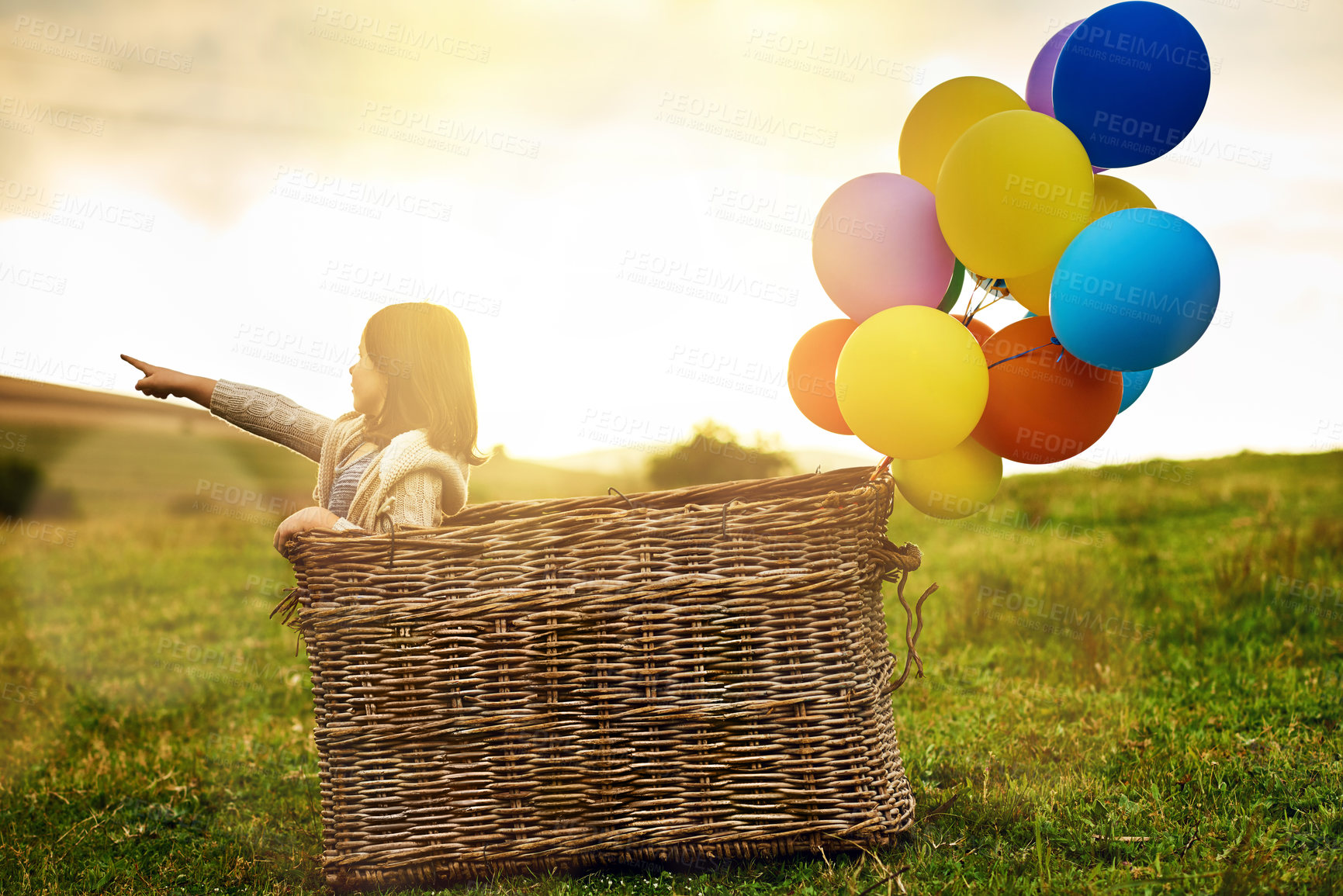 Buy stock photo Balloon, basket and girl with adventure in field for imagination, creativity or explore on holiday. Nature, kid and invention with point at sunset for fantasy travel direction, location and childhood