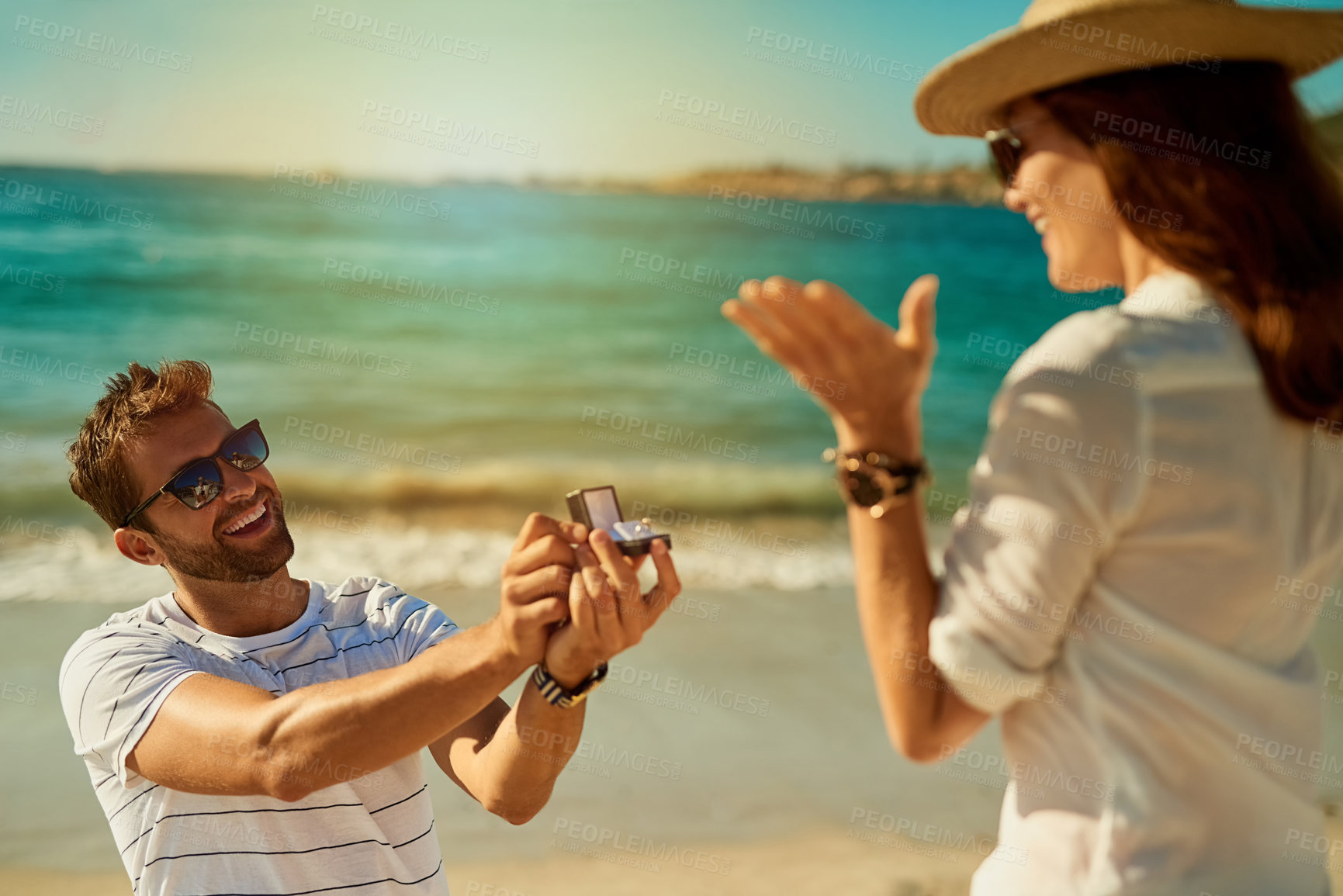Buy stock photo Shot of a young man proposing to his girlfriend at the beach on a summer’s day