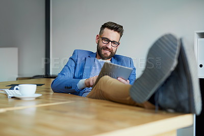 Buy stock photo Shot of a young businessman working on a digital tablet with his feet up on the desk in an office