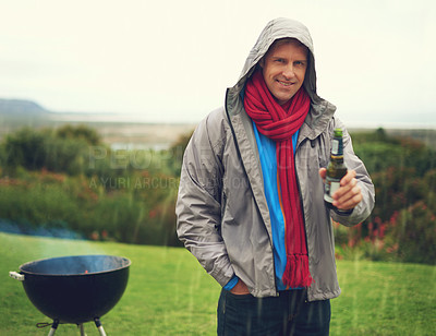 Buy stock photo Shot of a cheerful man barbecuing in the rain while drinking a beer