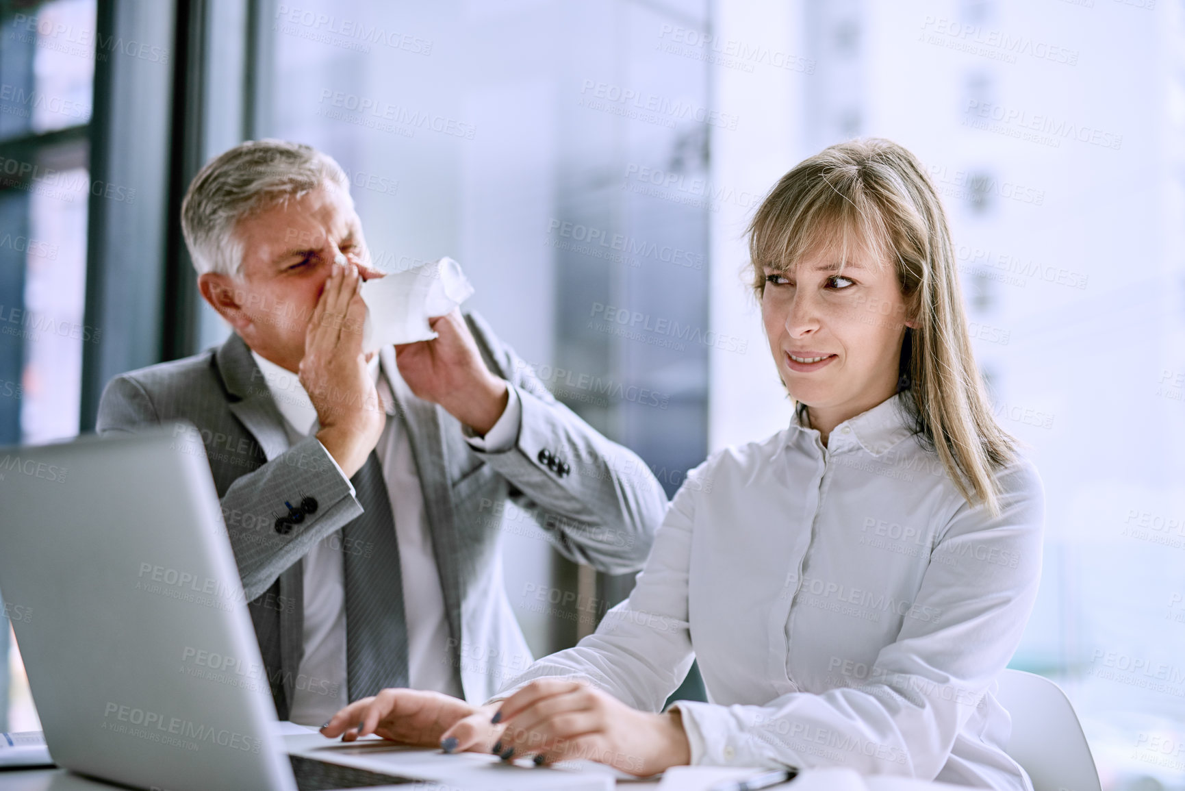 Buy stock photo Shot of a sick businessman sneezing in front of a businesswoman in the office