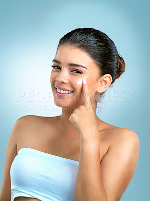 Buy stock photo Studio shot of an attractive  young woman applying moisturiser on her face against a blue background