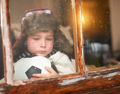 Buy stock photo Shot of a sad little boy holding a soccer ball while watching the rain through a window
