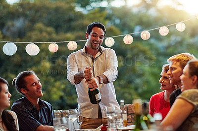 Buy stock photo Shot of a happy young man opening a bottle of champagne at a backyard party with friends