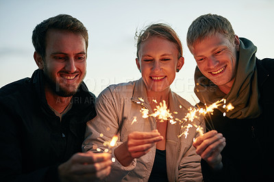 Buy stock photo Shot of a group of happy young friends playing with sparklers on the beach at sunset
