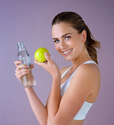 Buy stock photo Studio shot of a healthy young woman holding a glass water bottle and an apple against a purple background