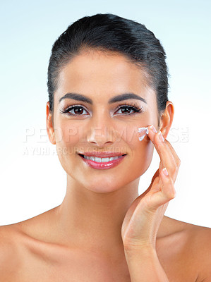 Buy stock photo Studio shot of a beautiful young woman applying moisturizer onto her face against a blue background