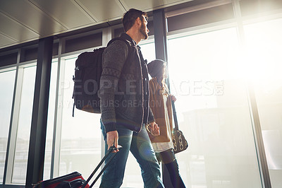 Buy stock photo Shot of a young couple walking inside of an airport with their luggage and holding hands while looking outside