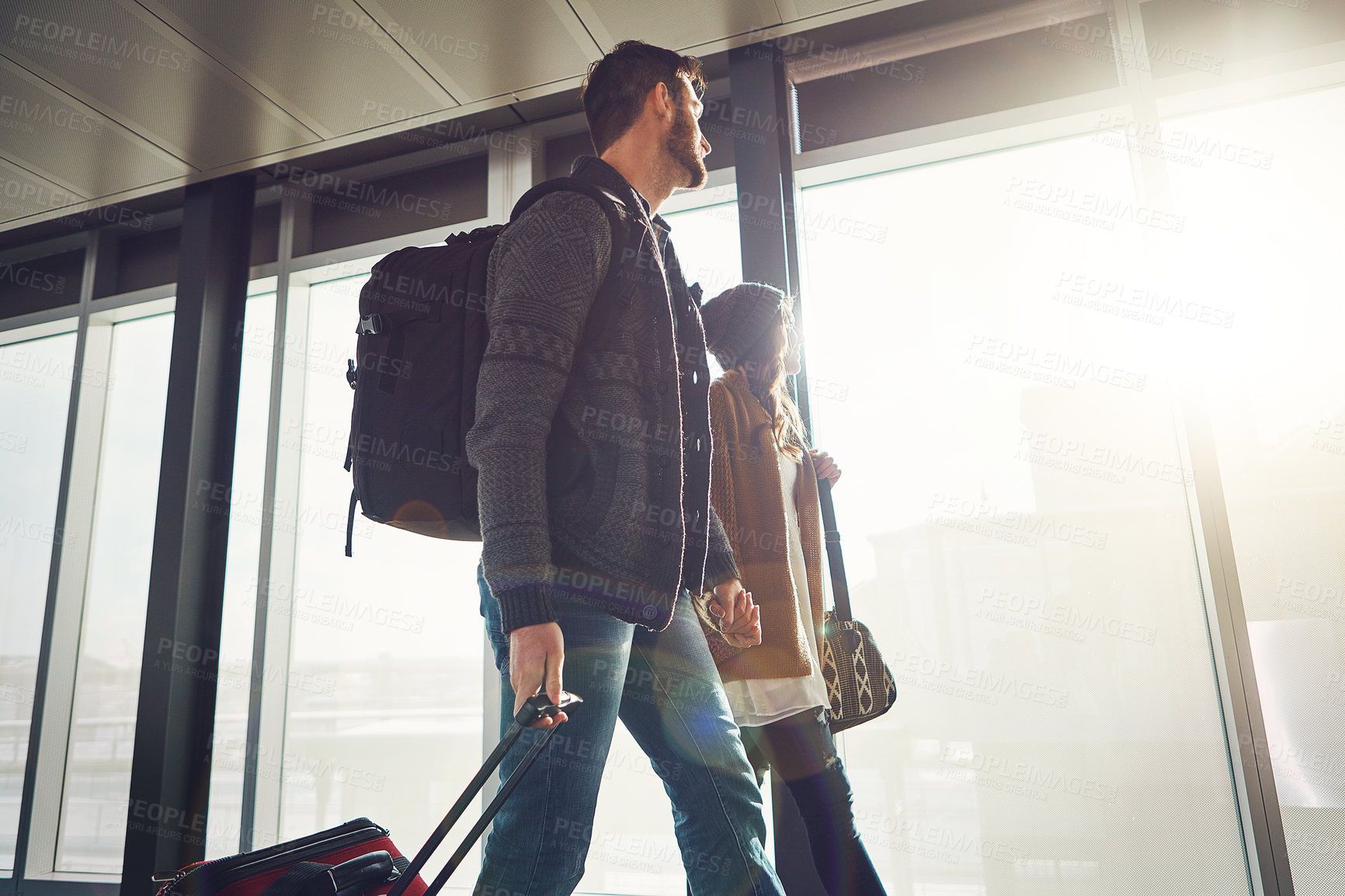 Buy stock photo Shot of a young couple walking inside of an airport with their luggage and holding hands while looking outside