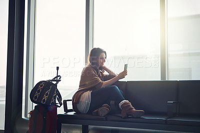 Buy stock photo Shot of a young woman sitting in an airport with her luggage and holding her cellphone and taking a picture