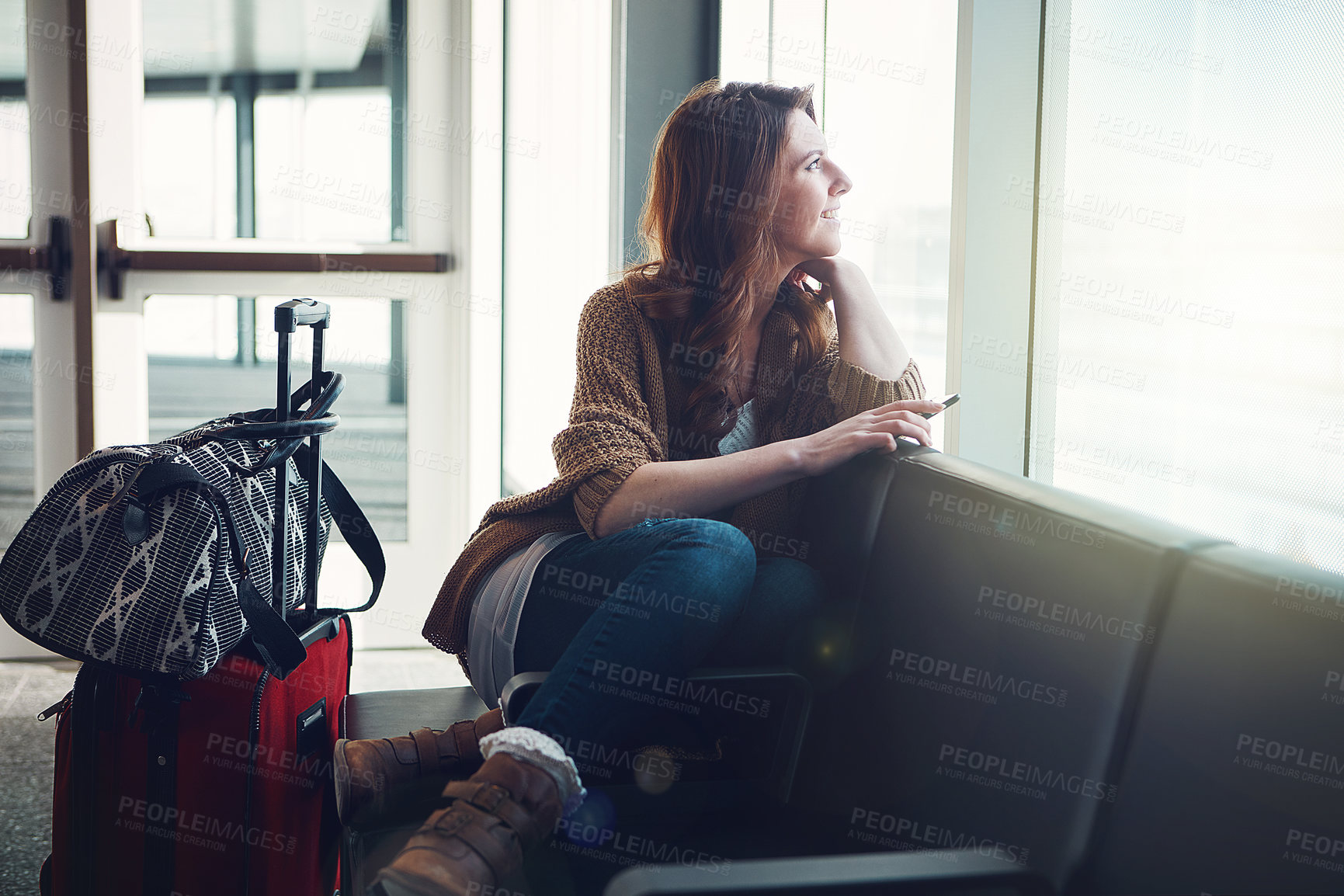 Buy stock photo Shot of a young woman sitting inside of an airport with her luggage and holding her cellphone while looking outside