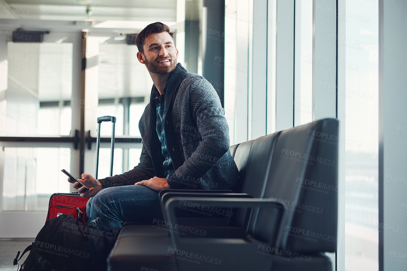 Buy stock photo Shot of a young man sitting in an airport with his luggage while holding his cellphone and looking at something