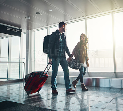 Buy stock photo Shot of a young couple walking in an airport with their luggage while holding hands and looking at one another