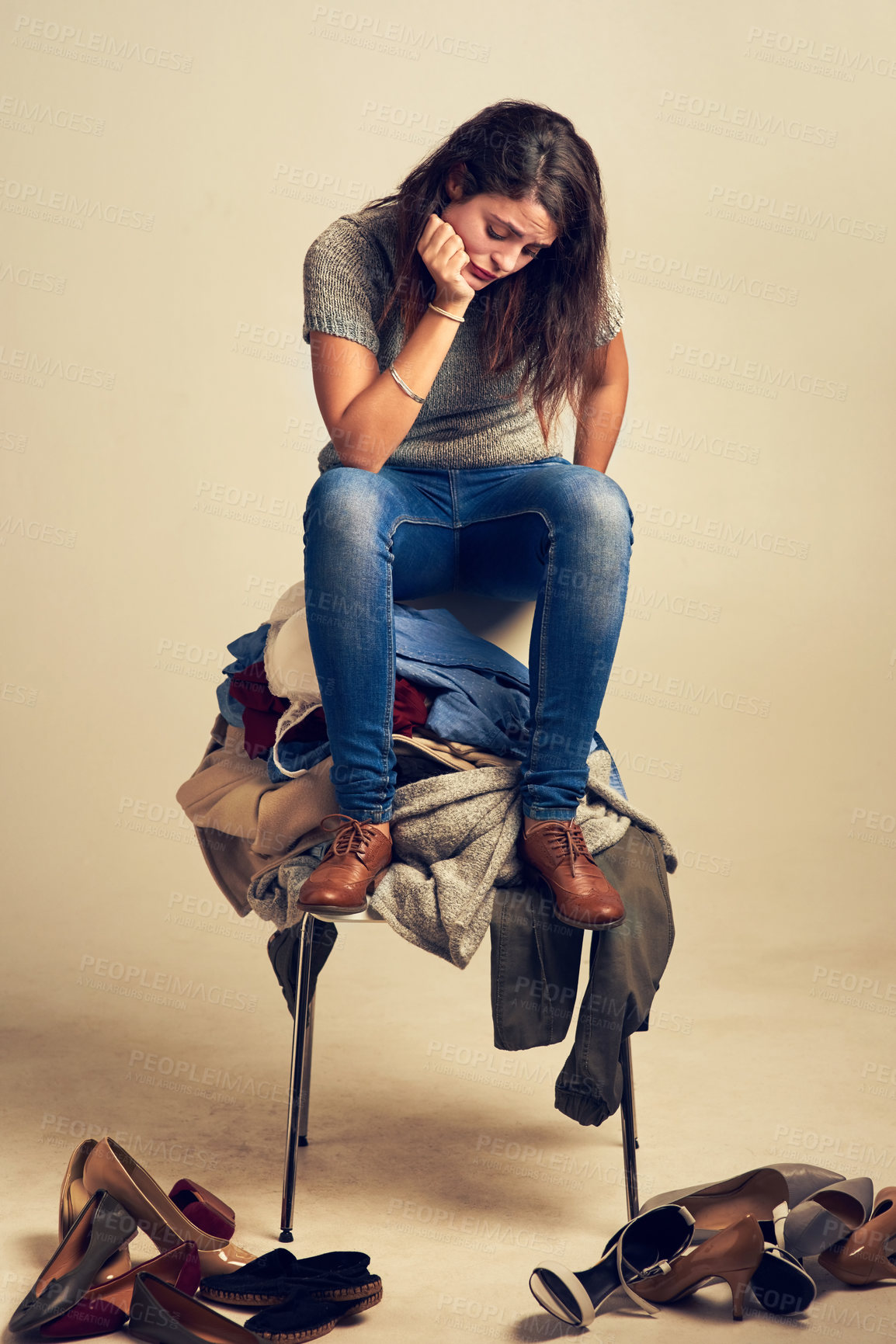 Buy stock photo Studio shot of a young woman trying to choose an outfit from a pile of clothes against a brown background