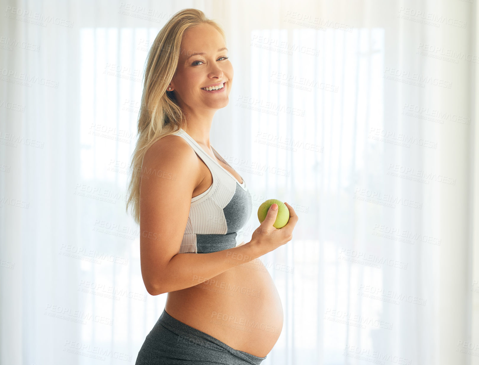 Buy stock photo Portrait of a happy pregnant woman dressed in sportswear and eating an apple at home