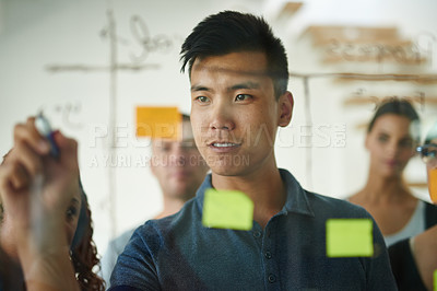Buy stock photo Closeup of marketing professional brainstorming ideas with colleagues, writing on transparent board during meeting. Portrait of young, Asian entrepreneur discussing work with employees in background.