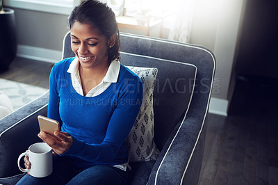 Buy stock photo Shot of a young woman using a mobile phone and having coffee on the sofa at home