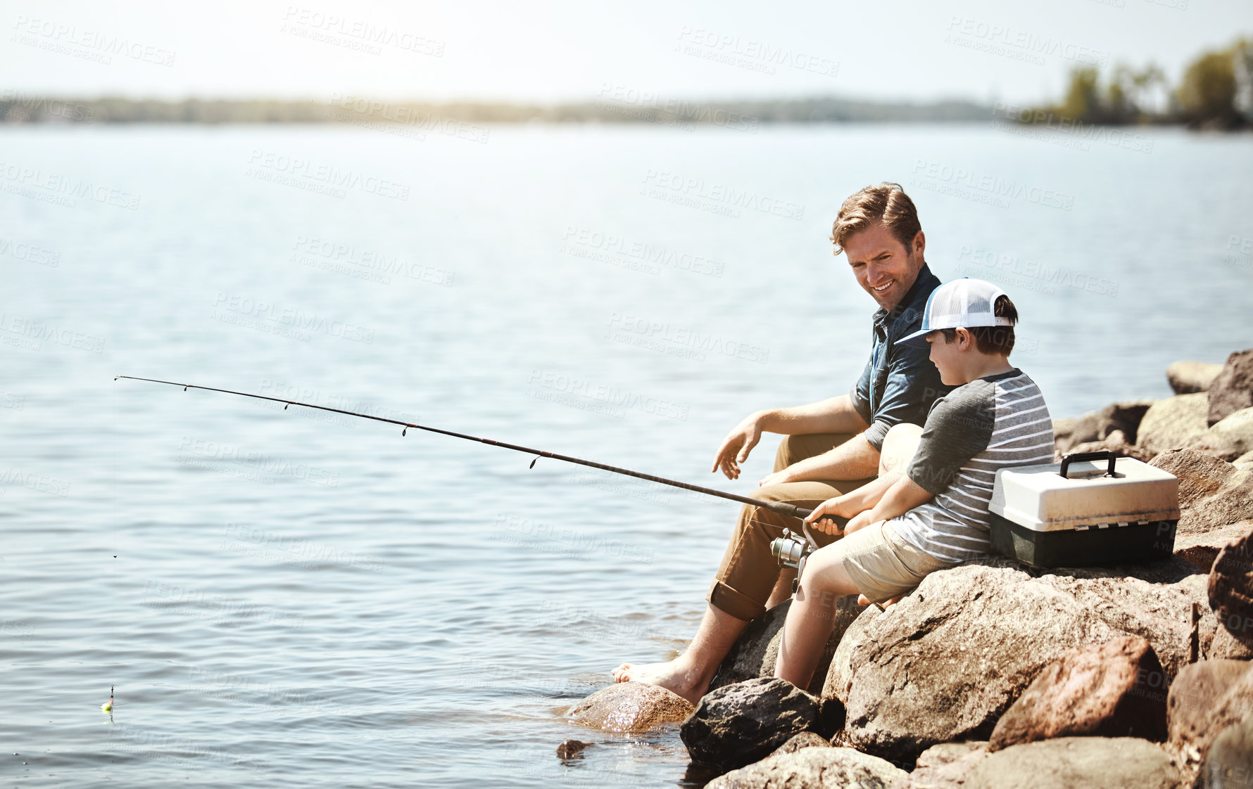 Buy stock photo Happy father, son and learning with fishing rod on rock by lake, ocean or beach together in nature. Dad with young child, kid or little boy for bonding, teaching tips or lesson to catch by sea water