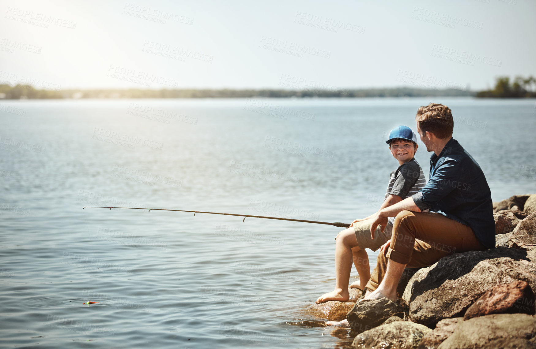 Buy stock photo Happy boy, father and learning with fishing rod on rock by lake, ocean or beach together in nature. Dad with son, young kid or little child for bonding, teaching tips or lesson to catch by sea water