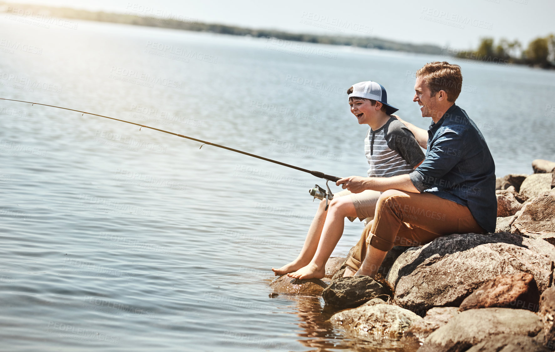 Buy stock photo Happy kid, father and learning with fishing rod on rock by lake, ocean or beach together in nature. Dad with son, young child or little boy for bonding, teaching tips or lesson to catch by sea water