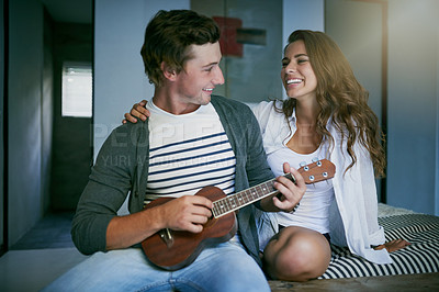 Buy stock photo Shot of a young man playing the ukulele while sitting on bed with his girlfriend