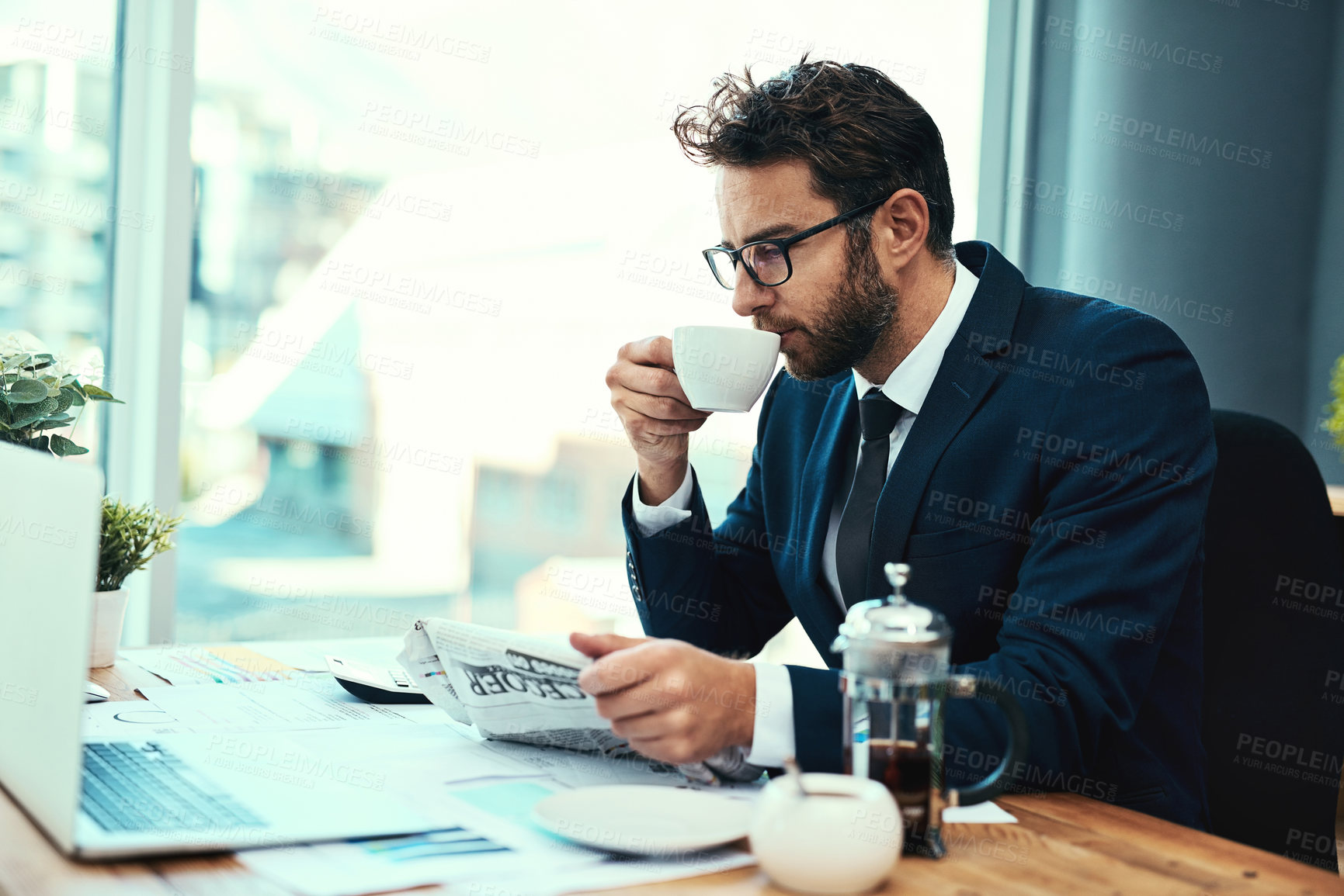 Buy stock photo Shot of a young businessman drinking a cup of tea while reading a newspaper in an office