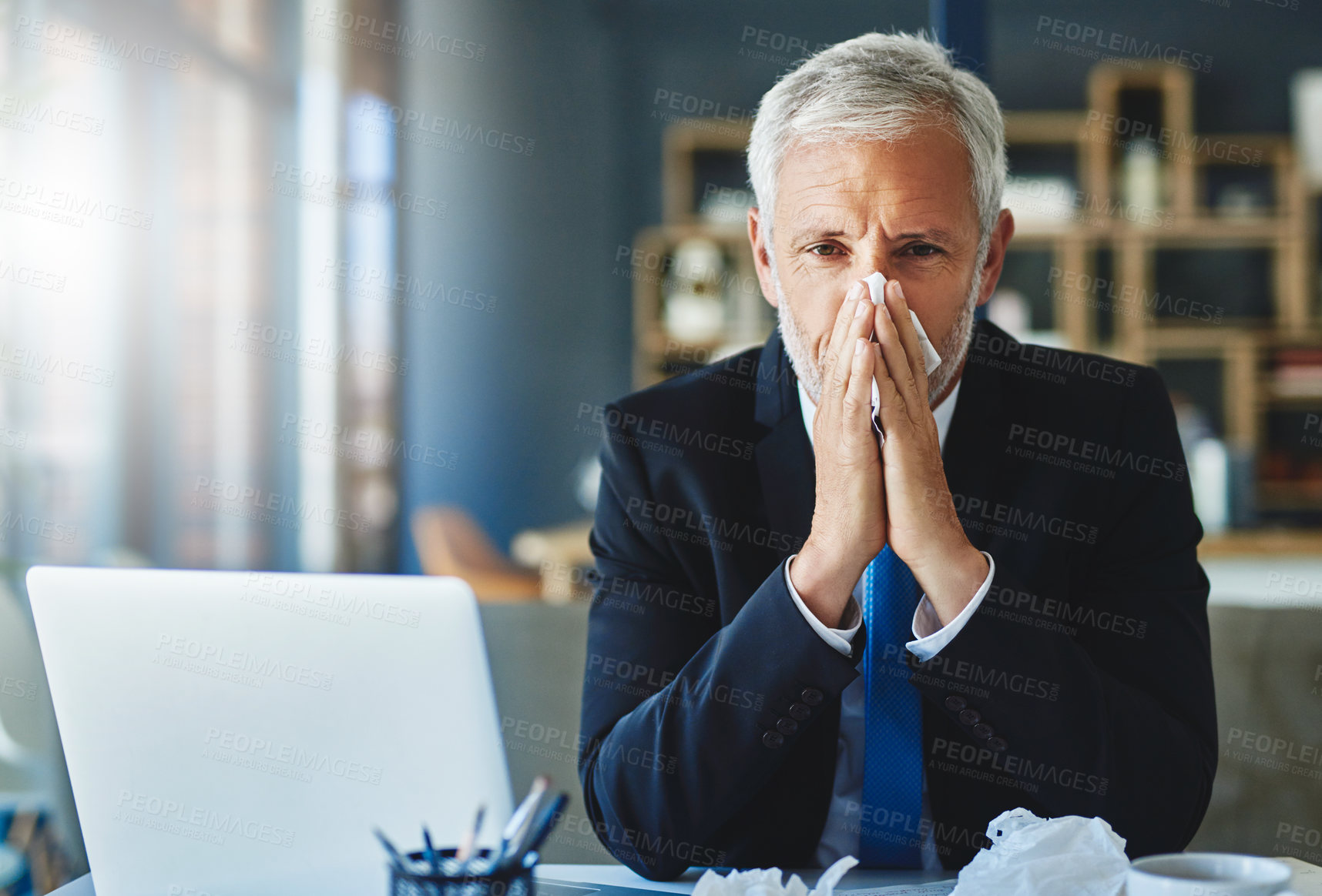 Buy stock photo Shot of a frustrated businessman using a tissue to sneeze in while being seated in the office