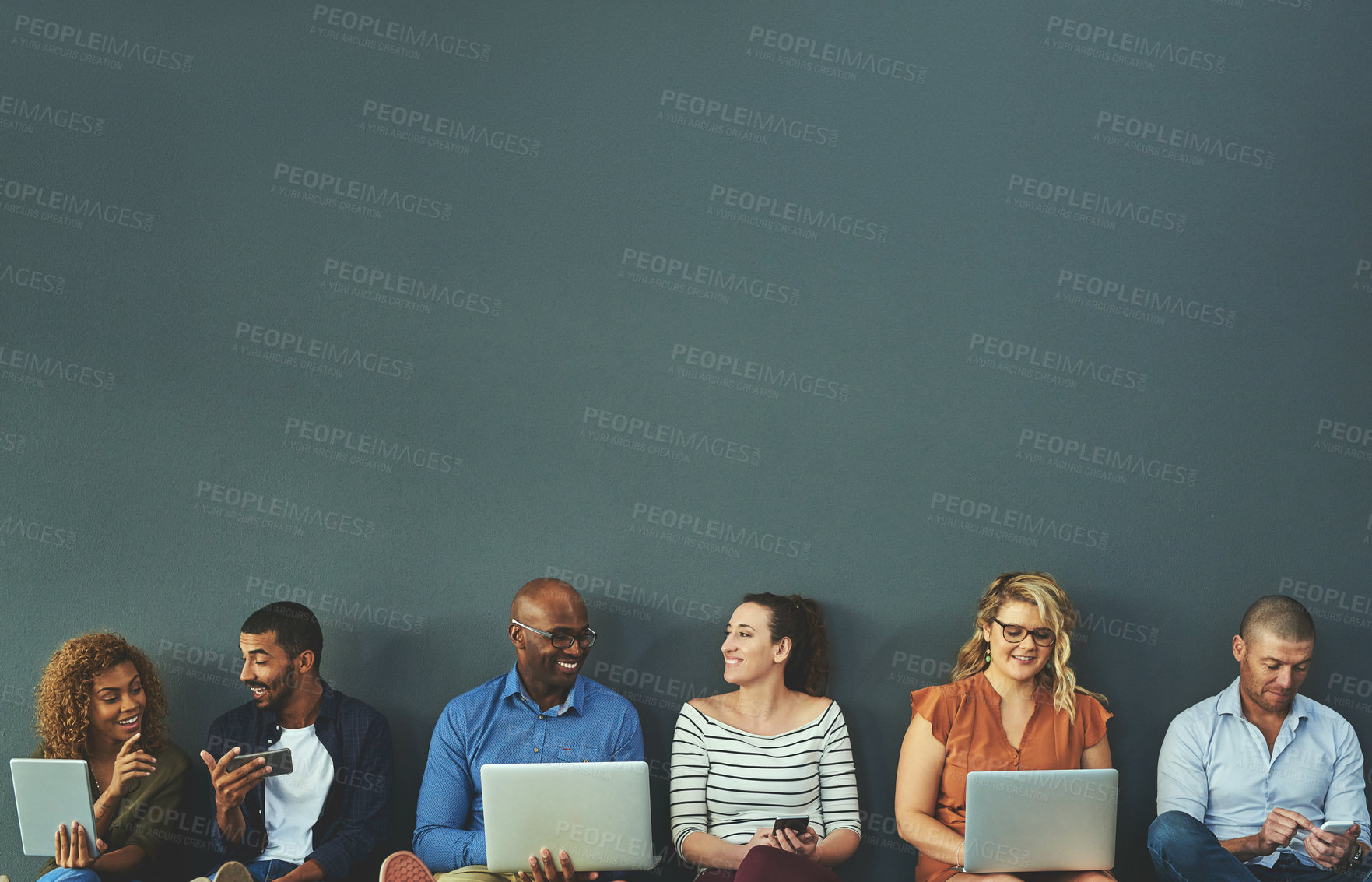 Buy stock photo Group of happy business people sharing information on wireless technology in a row at an office together. Corporate professionals sitting and talking using devices in a waiting room together at work