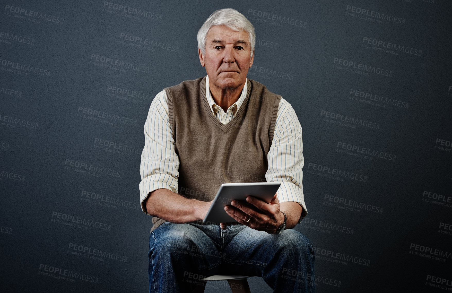 Buy stock photo Studio portrait of a handsome mature man using a tablet while sitting on a wooden stool against a dark background