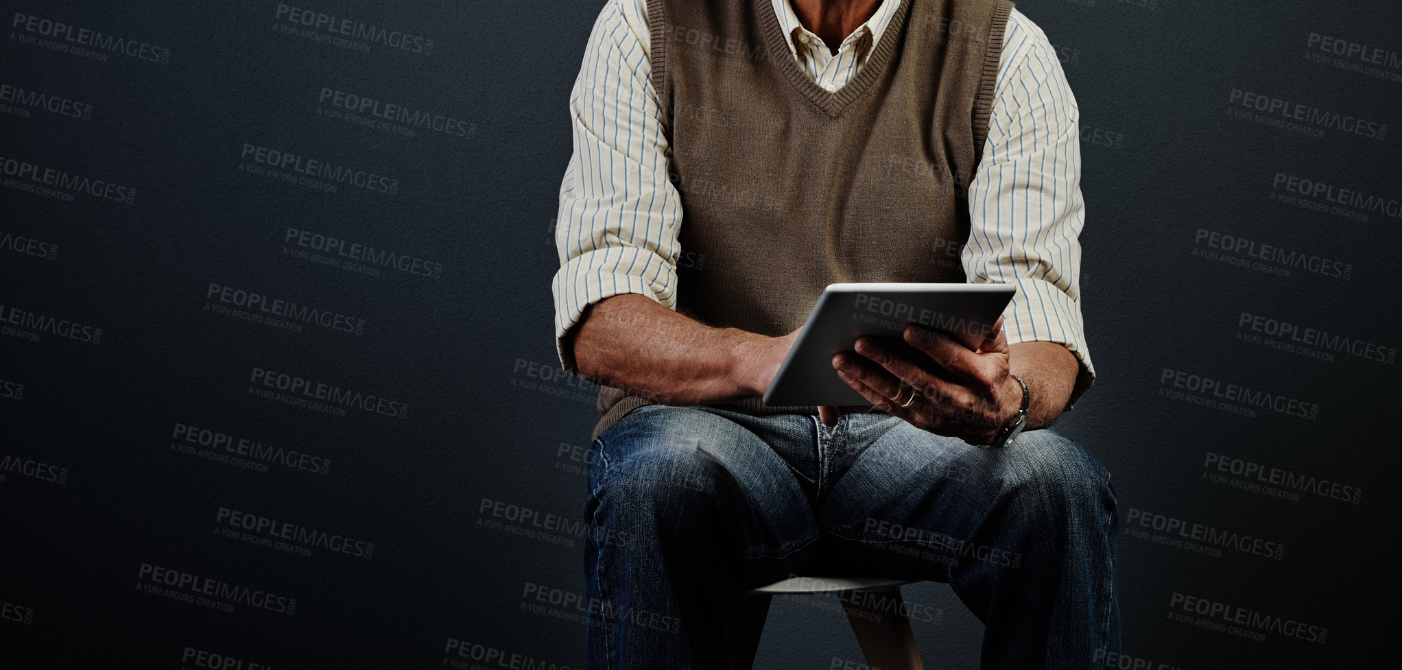 Buy stock photo Studio shot of an unrecognizable man using a tablet while sitting on a wooden stool against a dark background