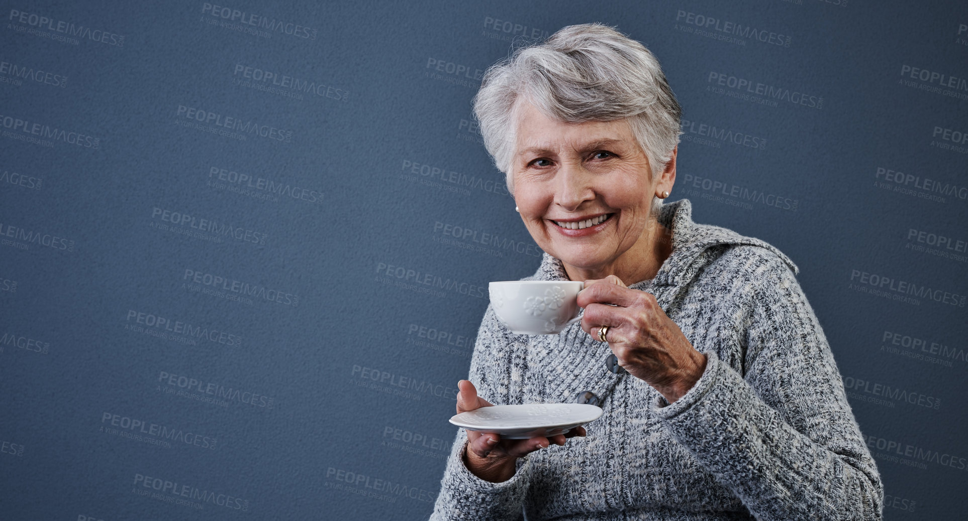 Buy stock photo Studio shot of a cheerful elderly woman sitting down and enjoying a cup of tea while looking at the camera