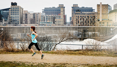 Buy stock photo Shot of a sporty young woman running alongside the city