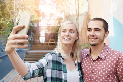 Buy stock photo Shot of a happy young couple using a mobile phone to take selfies outside