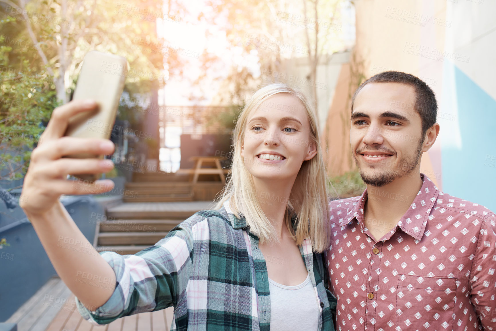 Buy stock photo Shot of a happy young couple using a mobile phone to take selfies outside