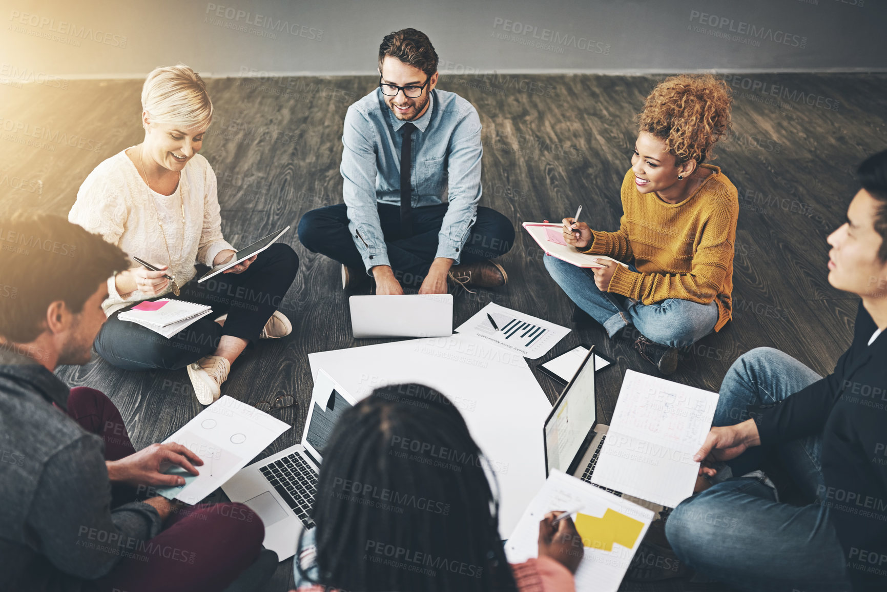 Buy stock photo Group of business people talking, planning with documents and sitting in a meeting on floor in an office together at work. Diverse, happy and smiling casual workers writing notes and browsing on tech