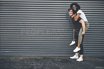 Buy stock photo Fun, trendy and happy interracial couple enjoying a piggyback ride together outside against an urban background. Lovers bonding and being playful. Smiling and relaxing while walking outdoors