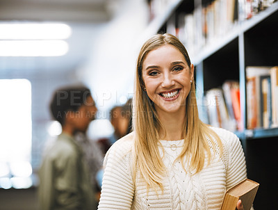 Buy stock photo Shot of a cheerful young woman holding a book while standing in a library and looking at the camera