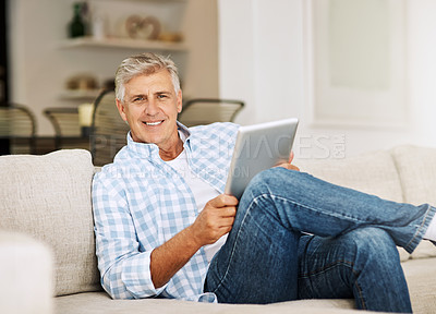 Buy stock photo Shot of a mature man using his digital tablet while relaxing on his sofa at home