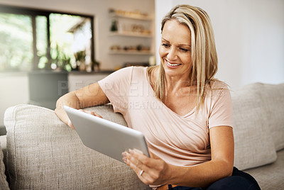 Buy stock photo Shot of a mature woman using her digital tablet while relaxing on her sofa at home