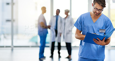 Buy stock photo Young male doctor reading, holding and looking at patient health information at a hospital. Medical professional standing with doctors in the background. Healthcare worker working on a clinic file