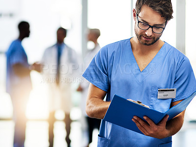 Buy stock photo Shot of a young doctor reading the contents of a file with his colleagues in the background