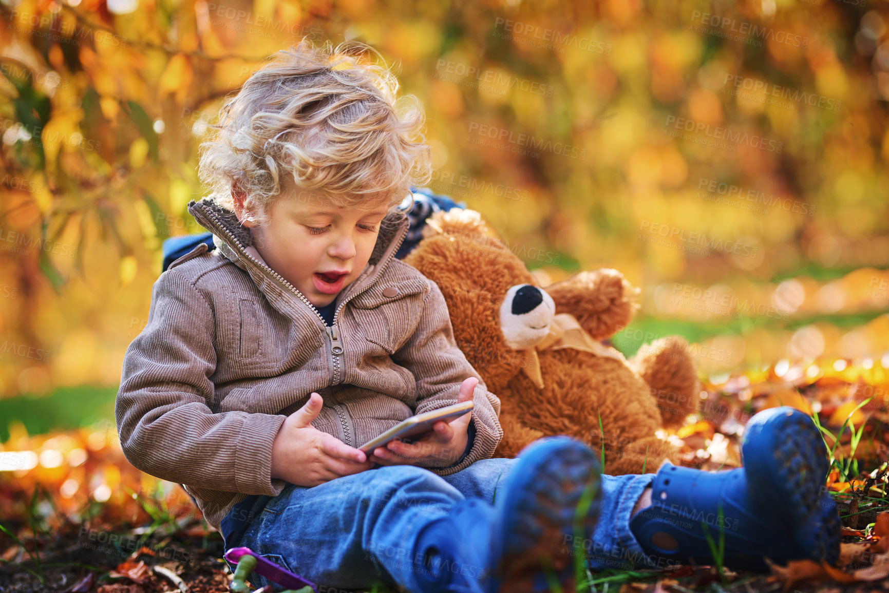 Buy stock photo Cropped shot of an adorable little boy using his cellphone while sitting outdoors during autumn