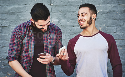 Buy stock photo Shot of two young men standing outdoors and using a mobile phone against a gray wall