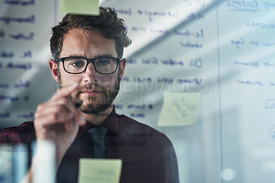 Buy stock photo Shot of a young businessman brainstorming with sticky notes on a glass wall in a modern office