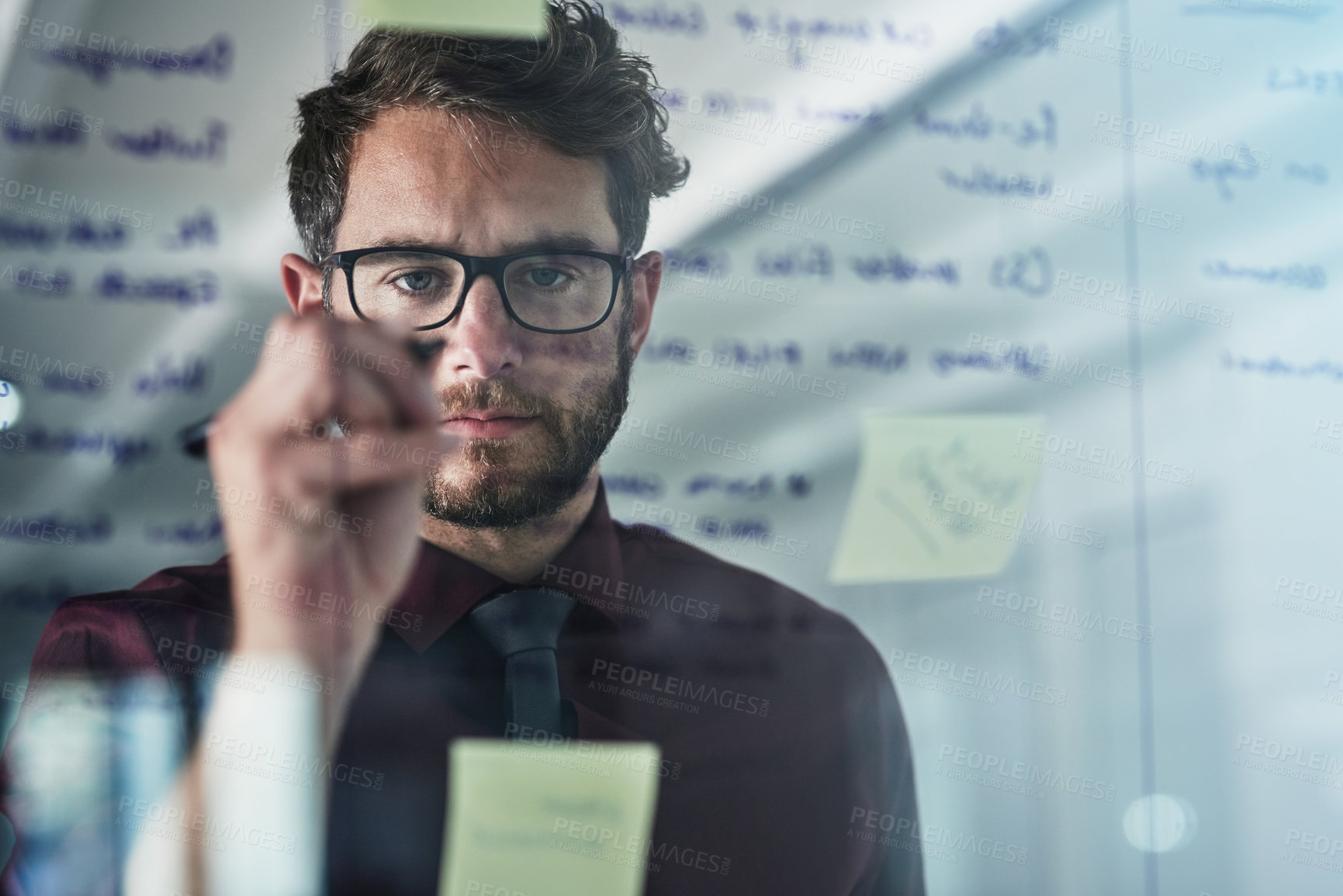 Buy stock photo Shot of a young businessman brainstorming with sticky notes on a glass wall in a modern office