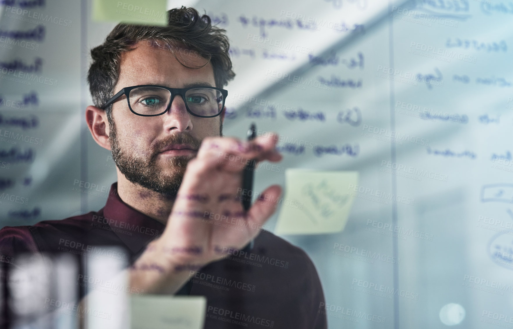 Buy stock photo Shot of a young businessman brainstorming with sticky notes on a glass wall in a modern office