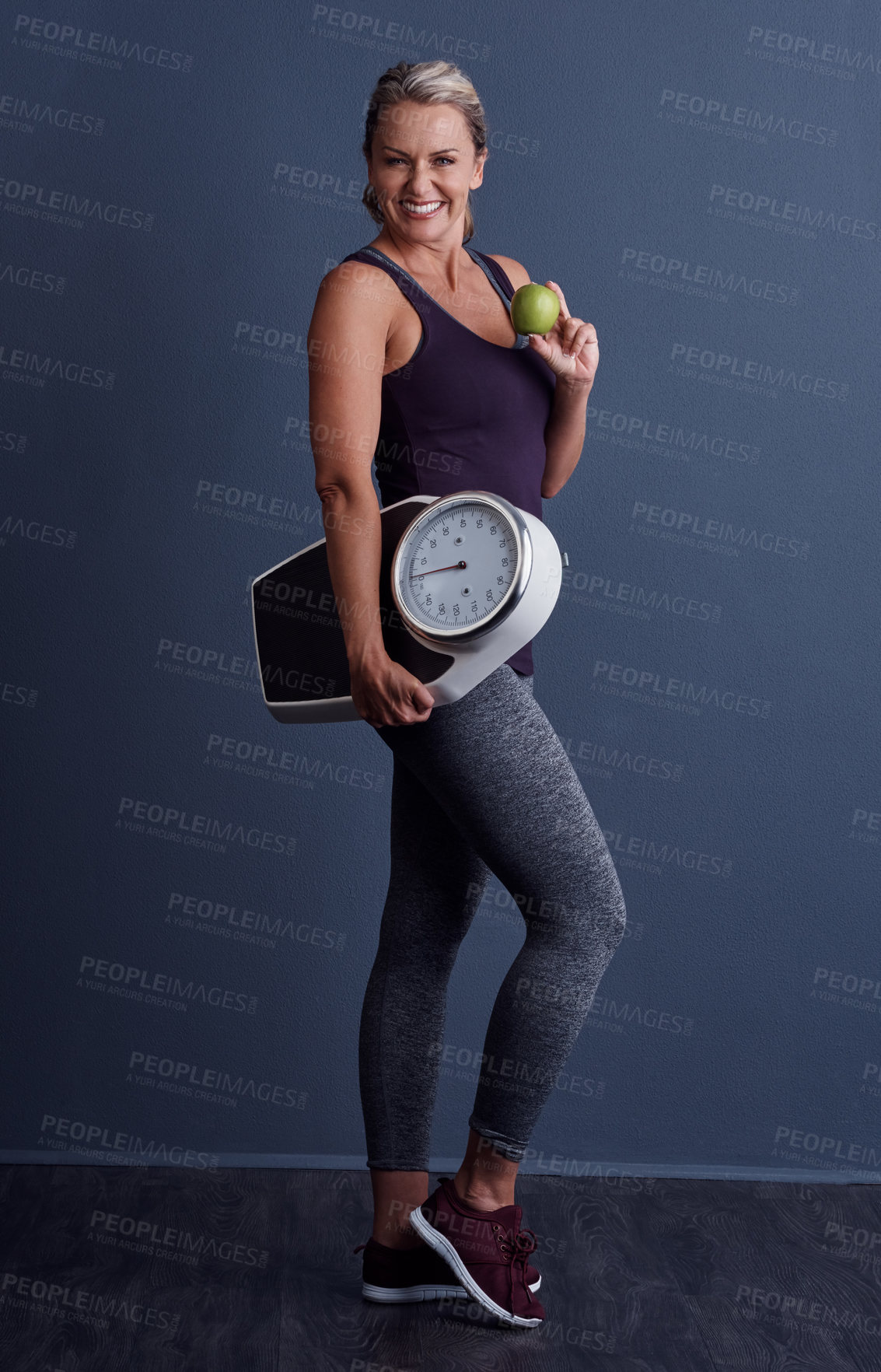 Buy stock photo Studio portrait of an attractive mature woman holding an apple and a weightscale against a blue background