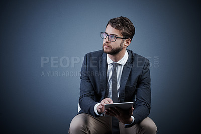 Buy stock photo Studio shot of a handsome young businessman using a tablet while sitting on a chair against a dark background