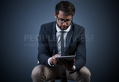 Buy stock photo Studio shot of a handsome young businessman using a tablet while sitting on a chair against a dark background