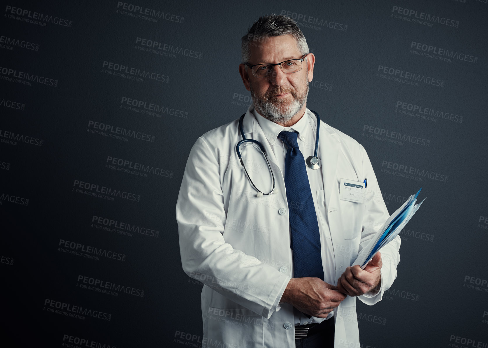 Buy stock photo Studio portrait of a handsome mature male doctor holding medical records while standing against a dark background